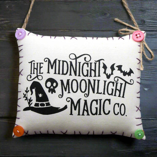Decorative pillow with 'The Midnight Moonlight Magic Co.' text and witch hat design on a wooden surface.