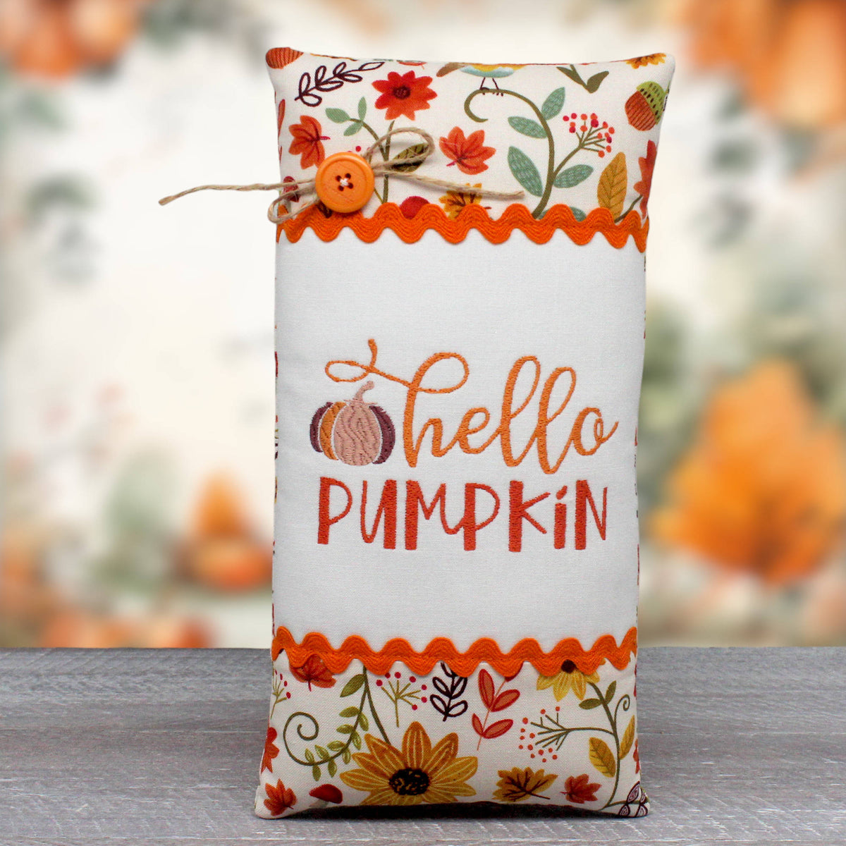 A decorative fabric pillow with an autumn 'Hello Pumpkin' design, featuring embroidered text, a pumpkin motif, and colorful floral patterns on a white background, complete with a twine bow and button closure.