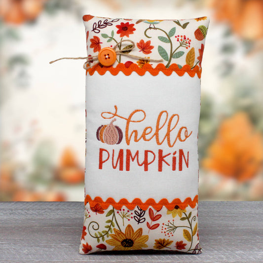 A decorative fabric pillow with an autumn 'Hello Pumpkin' design, featuring embroidered text, a pumpkin motif, and colorful floral patterns on a white background, complete with a twine bow and button closure.