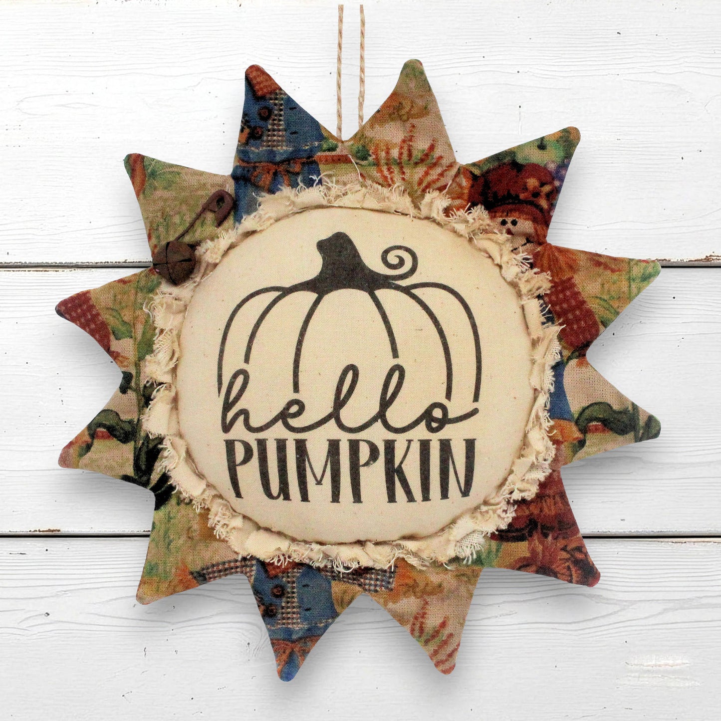 A sunflower-shaped fabric hanging ornament with a central cream circle that reads “hello PUMPKIN” beneath a black pumpkin outline. The surrounding star points are made from multicolored autumn-themed patterned fabric, including red, green, blue, and tan tones, and the ornament hangs against a white wood plank background.
