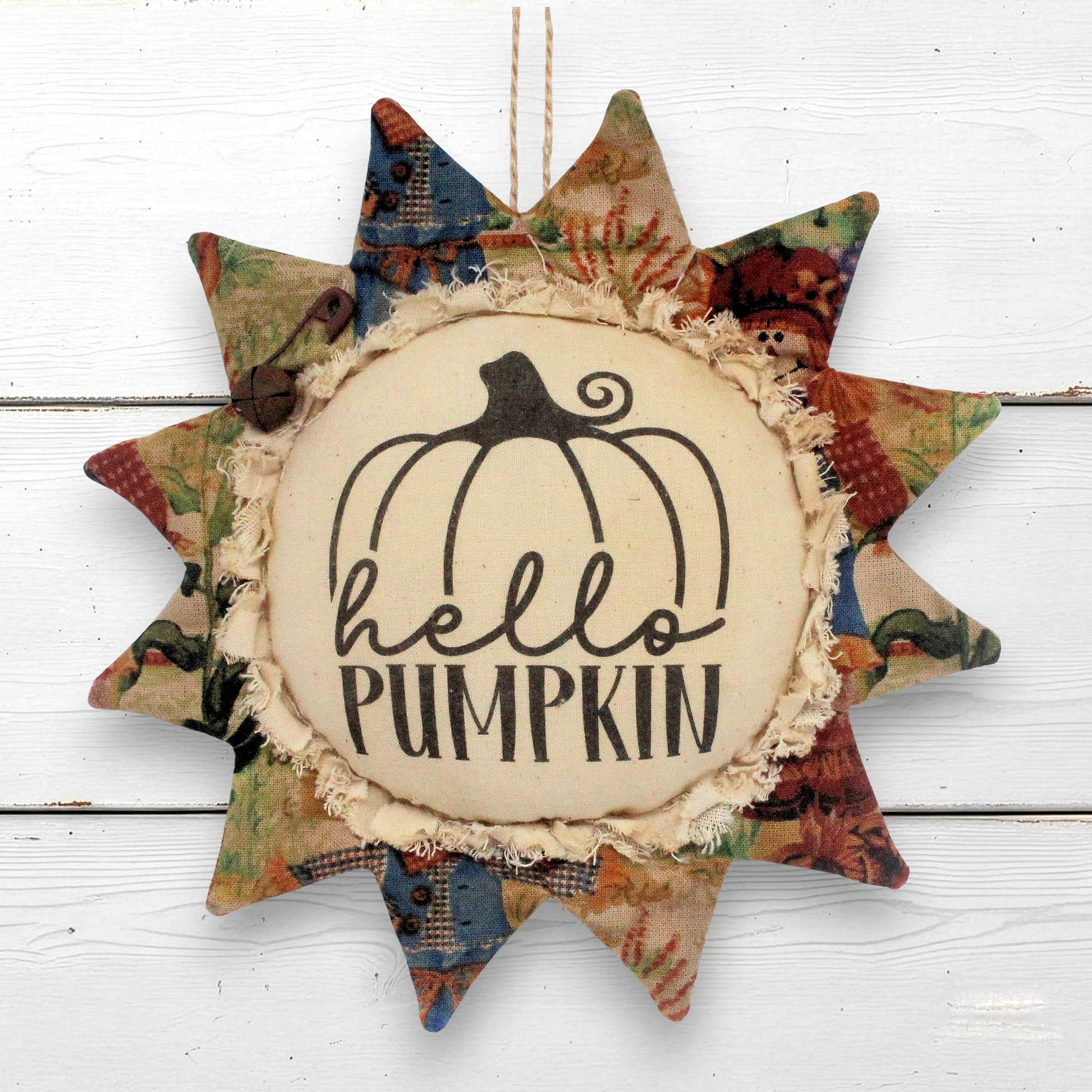 A sunflower-shaped fabric hanging ornament with a central cream circle that reads “hello PUMPKIN” beneath a black pumpkin outline. The surrounding star points are made from multicolored autumn-themed patterned fabric, including red, green, blue, and tan tones, and the ornament hangs against a white wood plank background.