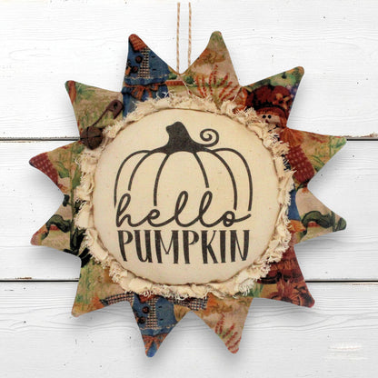 A sunflower-shaped fabric hanging ornament with a central cream circle that reads “hello PUMPKIN” beneath a black pumpkin outline. The surrounding star points are made from multicolored autumn-themed patterned fabric, including red, green, blue, and tan tones, and the ornament hangs against a white wood plank background.