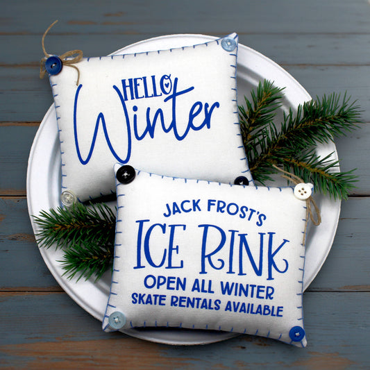 Two decorative mini pillow bowl fillers with winter-themed text on a white plate with greenery against a wooden background.