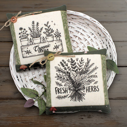 Set of two mini pillow bowl fillers with country primitive herb garden designs displayed on a white basket against a wooden background