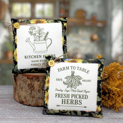 Two decorative fabric herb mini pillows styled on a rustic wood slice with dried yellow flowers. The pillows feature a black and yellow sunflower patterned border and a white center panel with black text reading 'KITCHEN HERBS' and 'FARM TO TABLE 100% ORGANIC FRESH PICKED HERBS'.