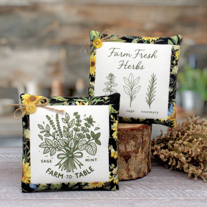 Two square fabric herb-themed mini pillows displayed on a rustic wooden surface. One pillow in front reads 'Farm to Table' with sage, mint, and rosemary illustrations, and the back pillow reads 'Farm Fresh Herbs' with botanical drawings.