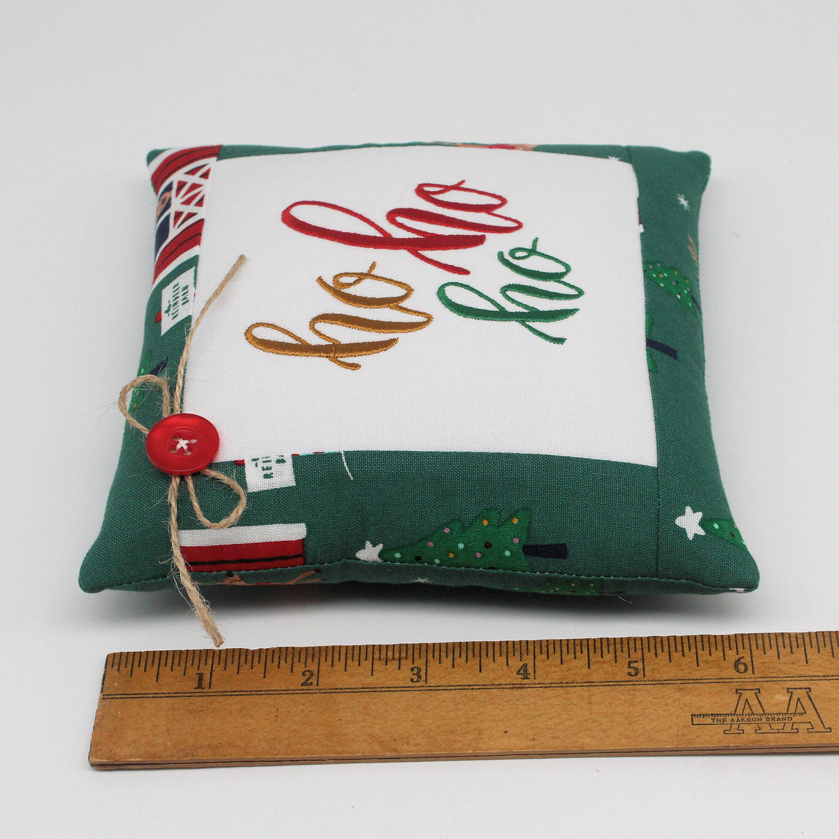 A small square fabric pillow with a green patterned border and a white center panel embroidered with 'ho ho ho' in red, gold, and green thread. A small red button and twine bow are attached on the left side.