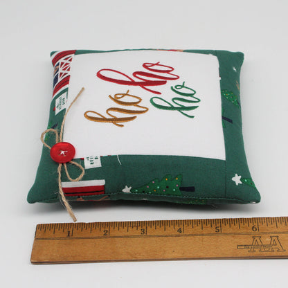 A small square fabric pillow with a green patterned border and a white center panel embroidered with 'ho ho ho' in red, gold, and green thread. A small red button and twine bow are attached on the left side.