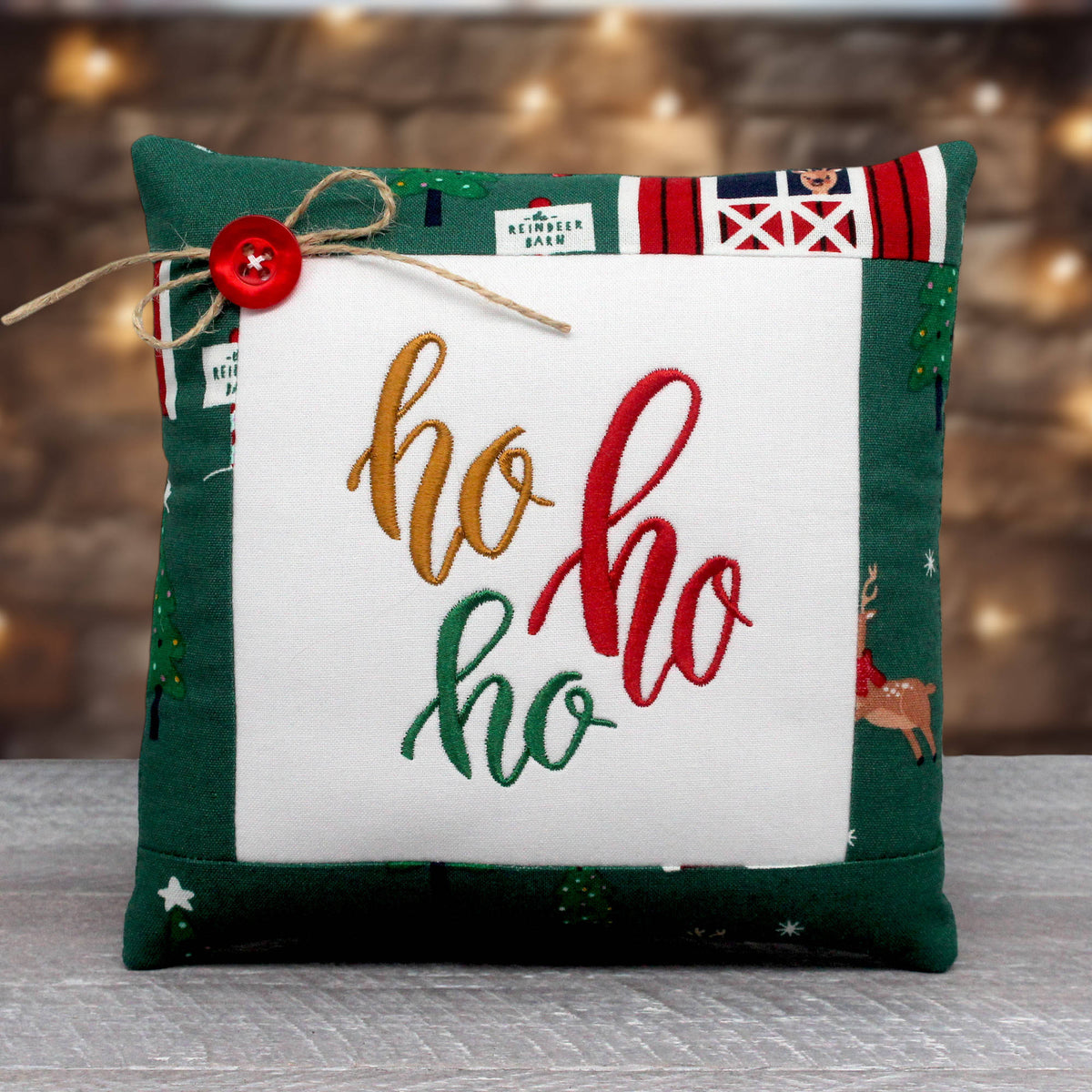 A square Christmas throw pillow with a green fabric border, a white center panel, and embroidered 'ho ho ho' text in gold, red, and green. The top edge features holiday-themed patchwork details, including a red button and twine bow, set against a blurred warm background.