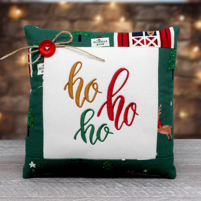 A square Christmas throw pillow with a green fabric border, a white center panel, and embroidered 'ho ho ho' text in gold, red, and green. The top edge features holiday-themed patchwork details, including a red button and twine bow, set against a blurred warm background.