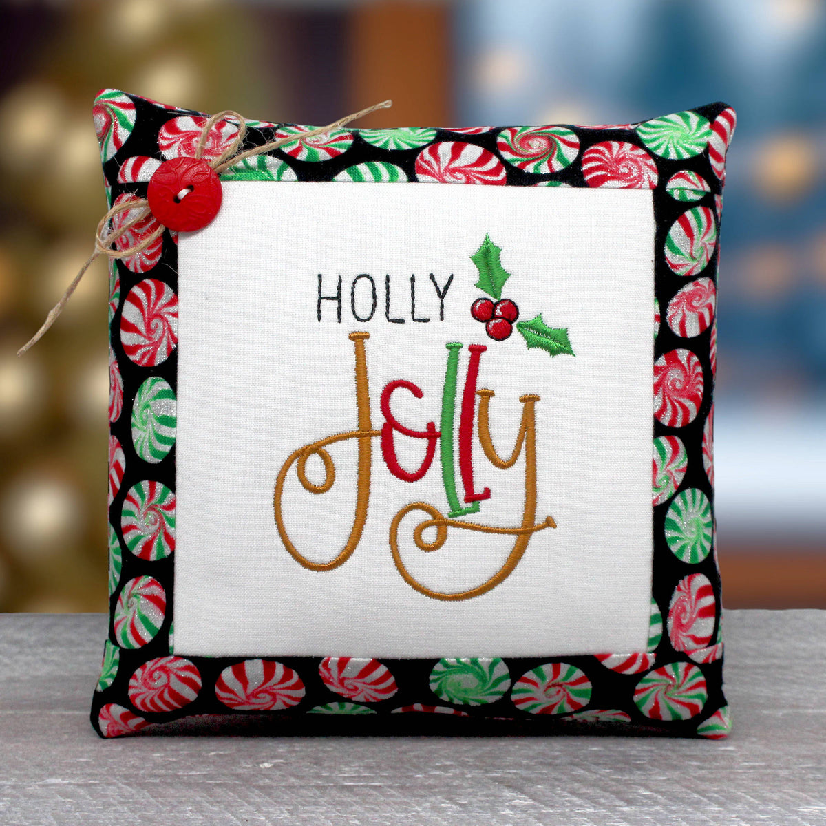 Small fabric decorative pillow tuck for Christmas with a machine embroidered design that reads "holly jolly."  It's trimmed with fabric that has a peppermint candy wheel design