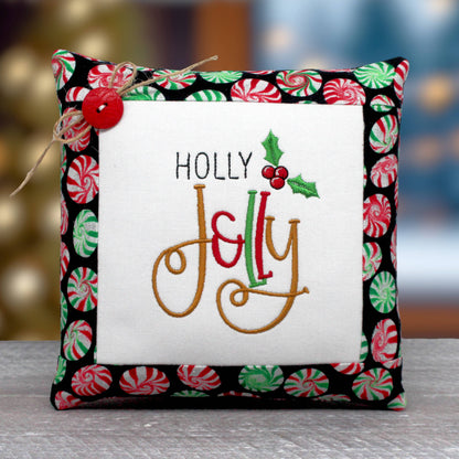 Small fabric decorative pillow tuck for Christmas with a machine embroidered design that reads "holly jolly."  It's trimmed with fabric that has a peppermint candy wheel design