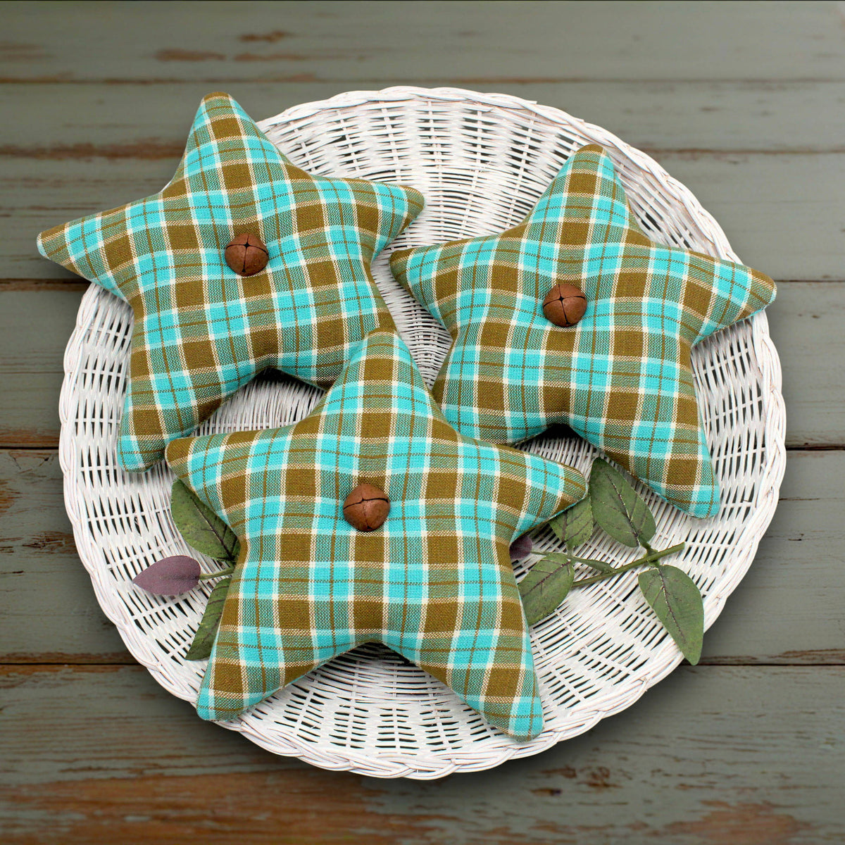 Three fabric stars in a green and turquoise plaid pattern with rusty bells, displayed on a white wicker tray over a rustic wooden surface.