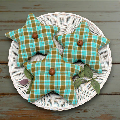 Three fabric stars in a green and turquoise plaid pattern with rusty bells, displayed on a white wicker tray over a rustic wooden surface.