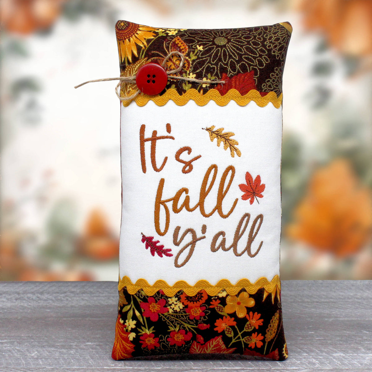 An elongated pillow tuck or bowl filler embroidered with 'It's Fall Y'all' and trimmed with rick rack and autumn-themed fabric