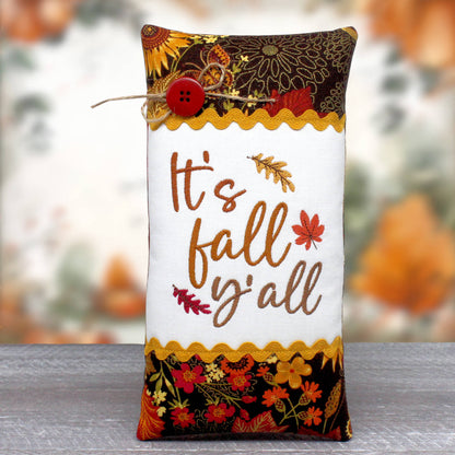 An elongated pillow tuck or bowl filler embroidered with 'It's Fall Y'all' and trimmed with rick rack and autumn-themed fabric