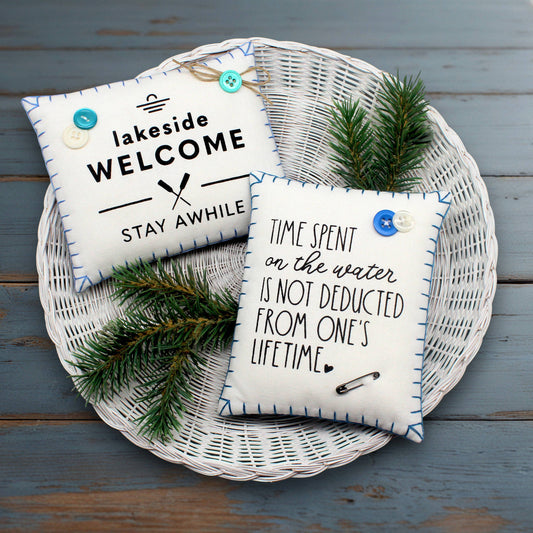 Two decorative pillows with lake quotes on a woven basket with greenery against a wooden background