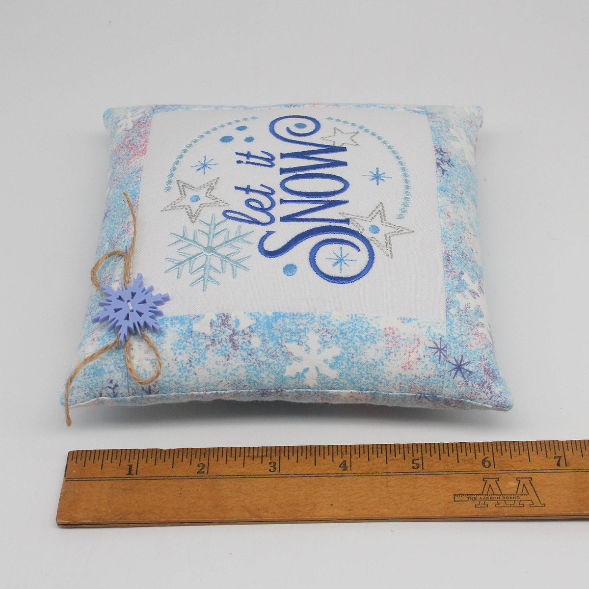 A square fabric pillow with a winter theme, featuring the embroidered text 'let it SNOW' in blue and silver, surrounded by snowflakes and stars. The pillow has a light blue and white patterned border, a small purple snowflake charm on a twine string, and is shown next to a wooden ruler for scale on a white background.