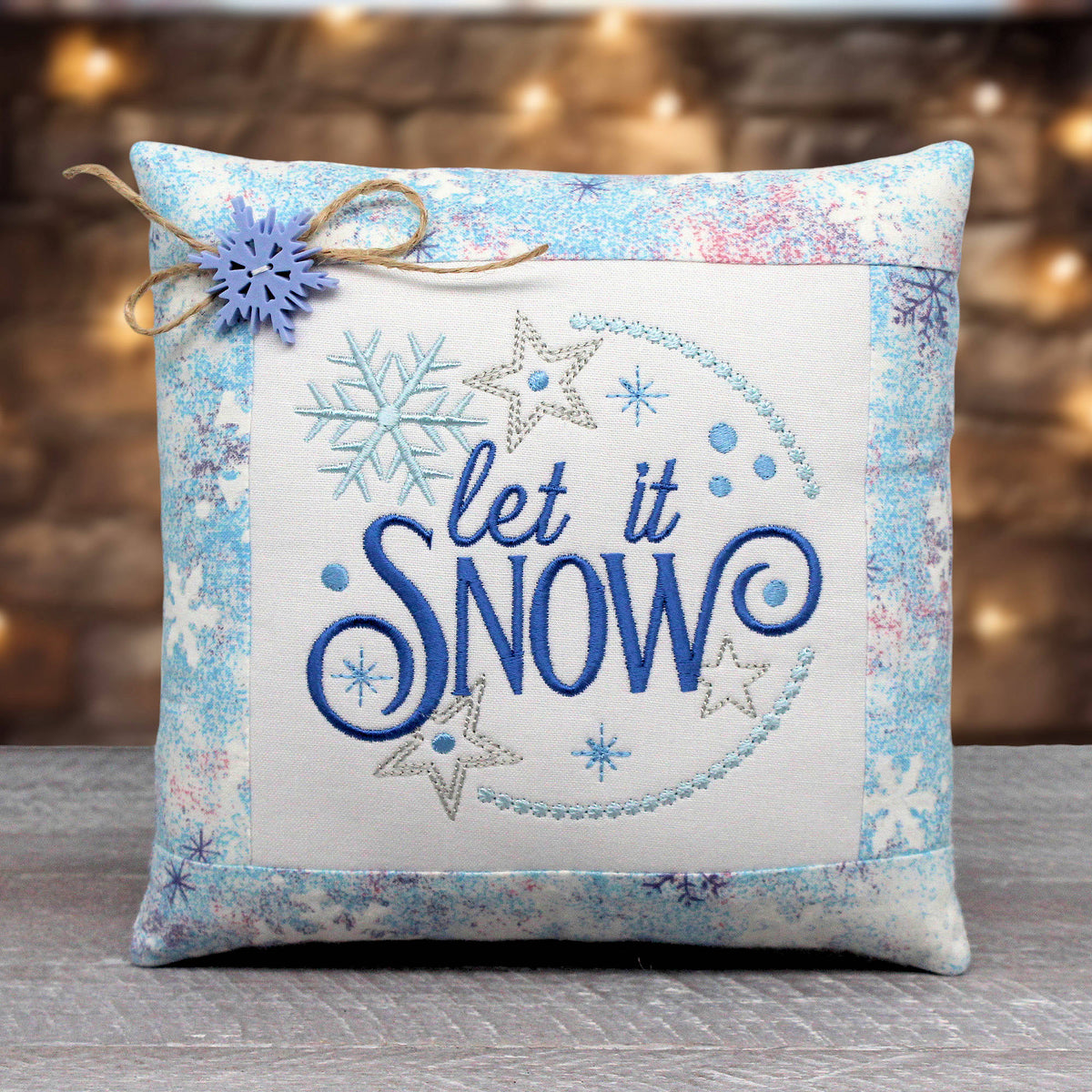 A square decorative throw pillow with a winter theme, featuring the embroidered phrase 'let it SNOW' in blue and silver on a cream center panel, surrounded by a light blue and white patterned border. A small blue snowflake ornament tied with twine is attached to the top left corner.
