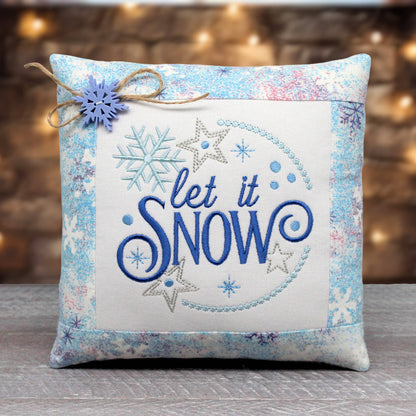 A square decorative throw pillow with a winter theme, featuring the embroidered phrase 'let it SNOW' in blue and silver on a cream center panel, surrounded by a light blue and white patterned border. A small blue snowflake ornament tied with twine is attached to the top left corner.