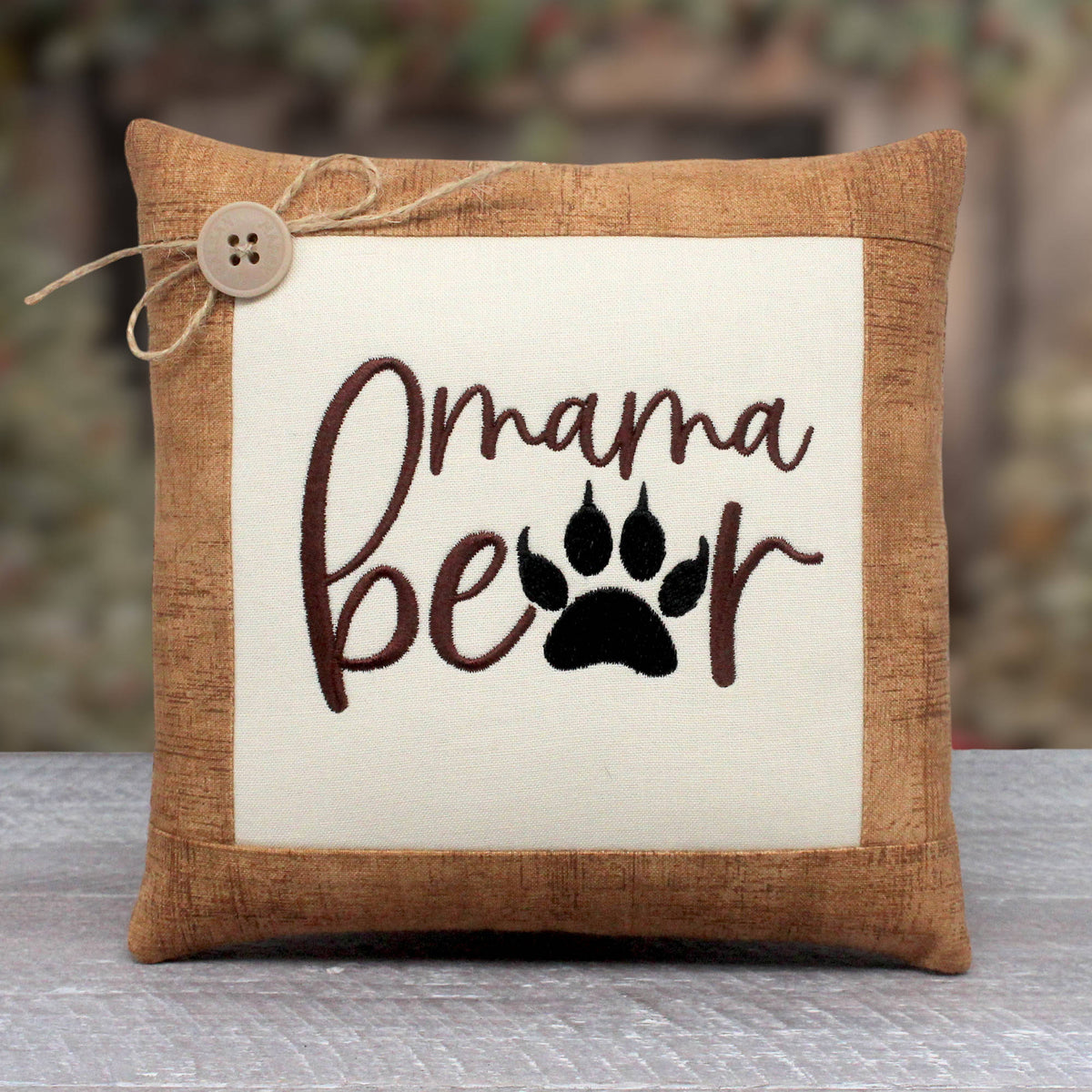 Small pillow tuck or bowl filler embroidered with the words 'mama bear' with a bear paw replacing the A in the word bear.  The center panel is trimmed with a light brown cotton print fabric.