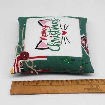 A small square Christmas-themed fabric pillow with a green border, a white center panel, embroidered cat ears and the words 'Meowy Christmas,' and a red button with twine closure. A wooden ruler is placed below the pillow to show scale.