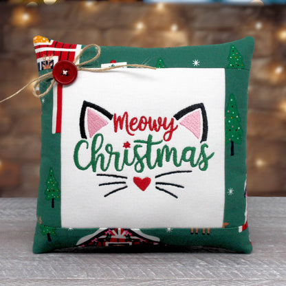 A square Christmas-themed decorative pillow with a white center panel featuring embroidered 'Meowy Christmas' text, cat ears, whiskers, and a red heart. The pillow has a dark green border with small Christmas tree and cabin motifs, plus a red button and twine bow on the left side.