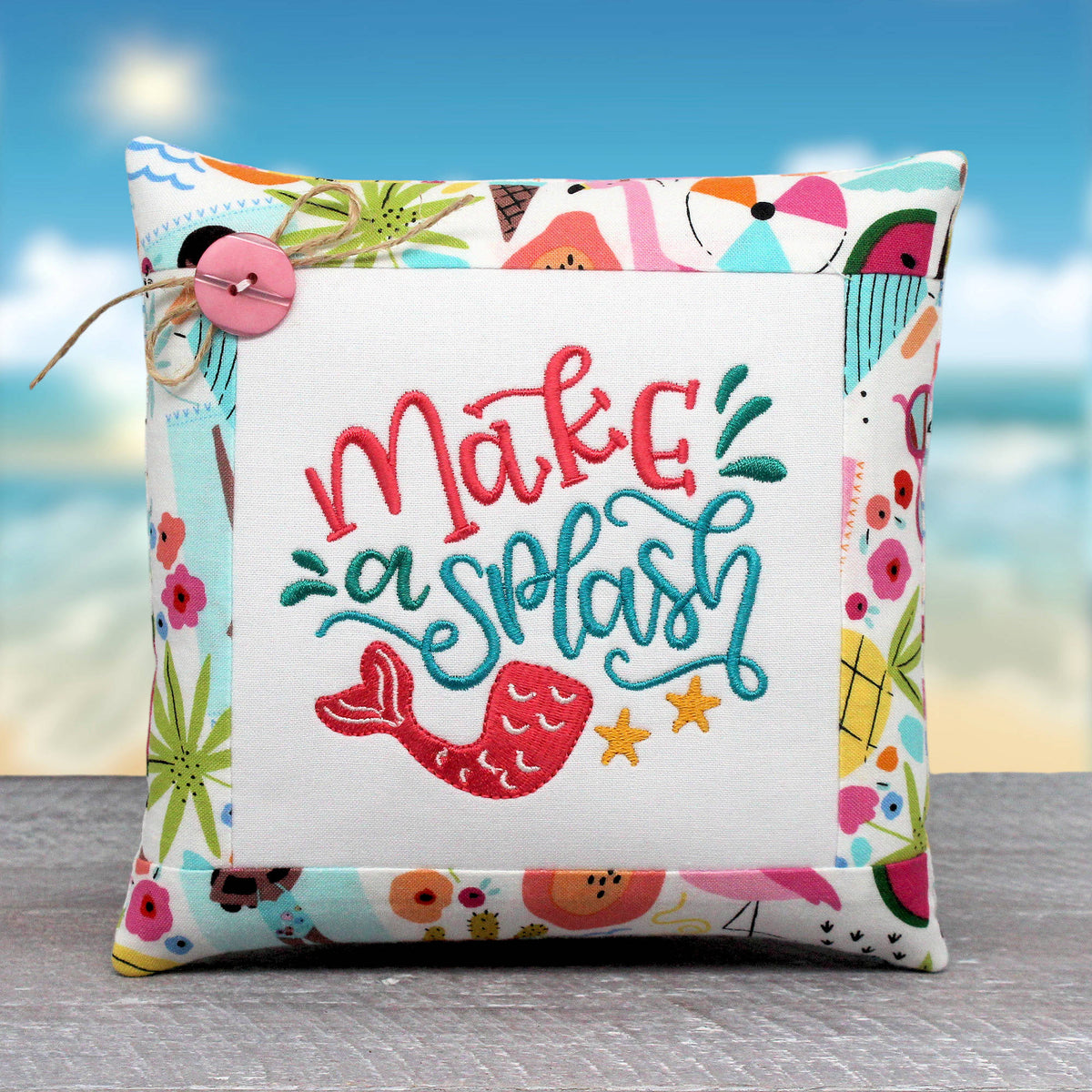 A square decorative throw pillow with a white center panel embroidered with the phrase 'make a splash' in coral and teal lettering, plus a mermaid tail and yellow starfish. The pillow has a colorful beach-themed border and is photographed against a blurred beach background.