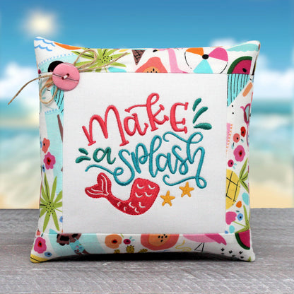 A square decorative throw pillow with a white center panel embroidered with the phrase 'make a splash' in coral and teal lettering, plus a mermaid tail and yellow starfish. The pillow has a colorful beach-themed border and is photographed against a blurred beach background.
