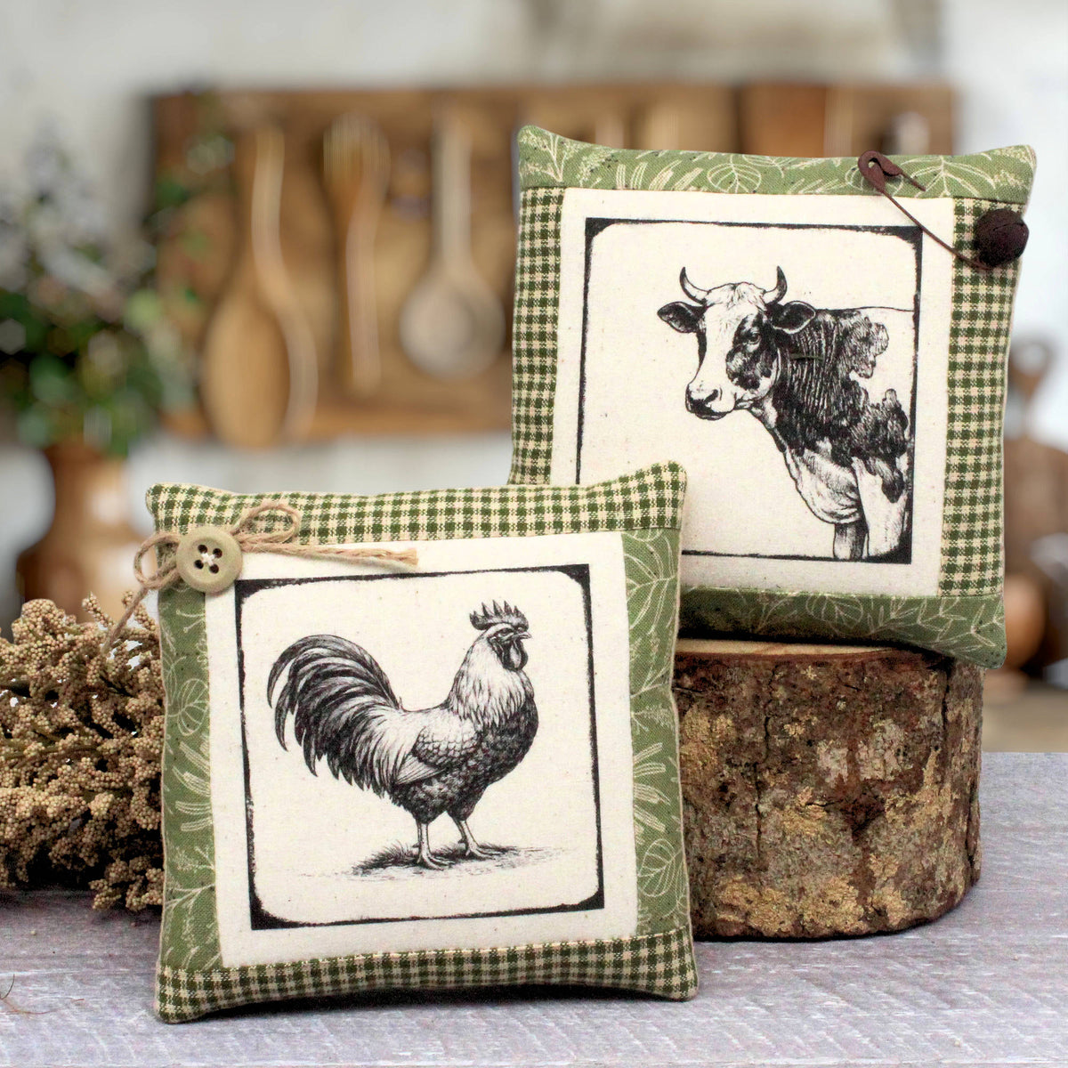 Two decorative square mini pillows with a rustic farmhouse design, featuring black and white animal illustrations (a rooster and a cow) on an off-white background with green gingham and leaf-patterned borders. One pillow is in the foreground and the other is stacked behind it on a rustic wood slice.