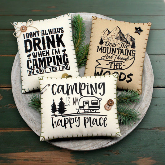 Three camping-themed pillows on a wooden surface with pine branches.