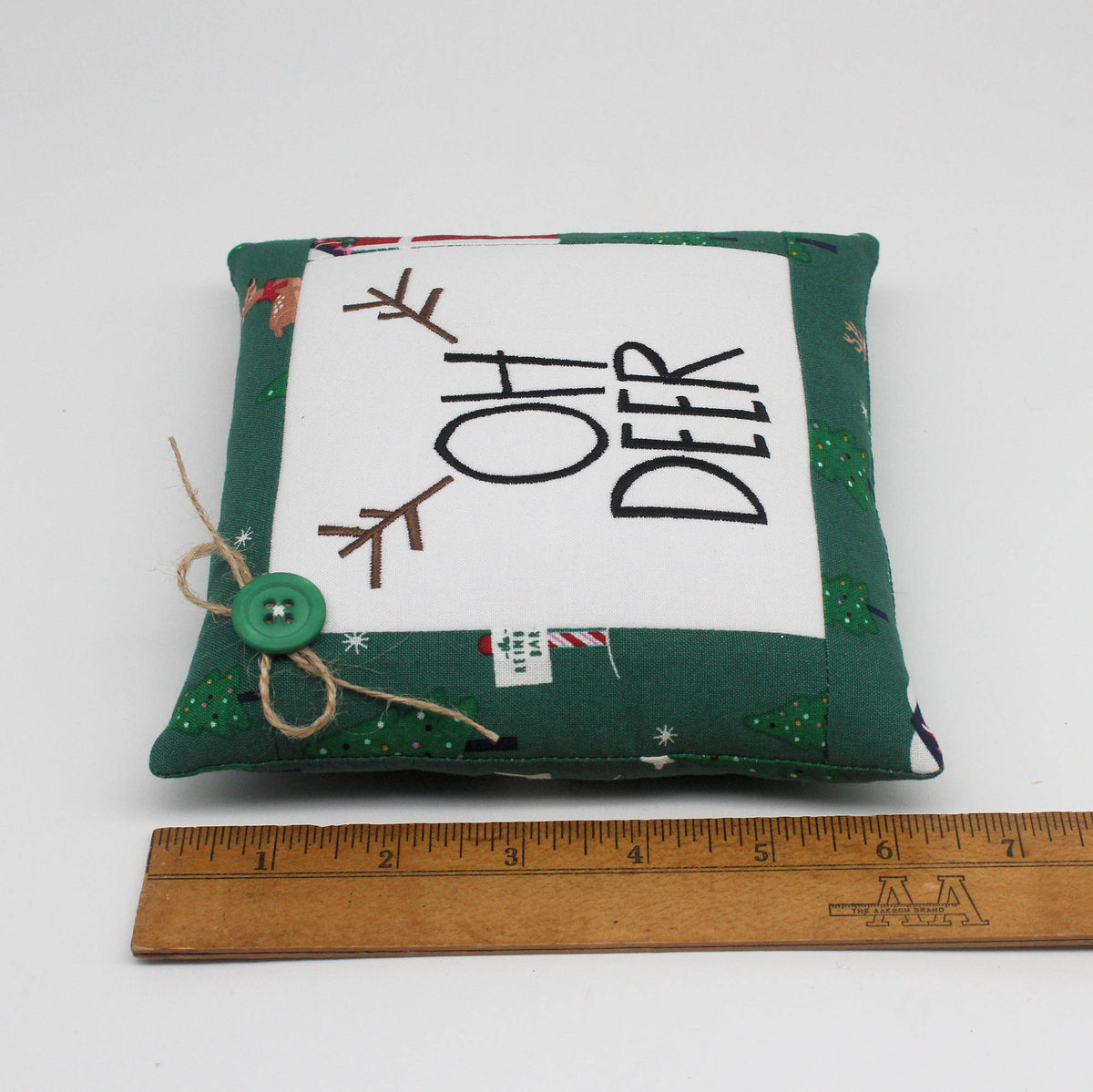 A square handmade fabric pillow with a green Christmas-themed border and a white center panel reading 'OH DEER' in black text. A wooden ruler is placed below the pillow to show its size.