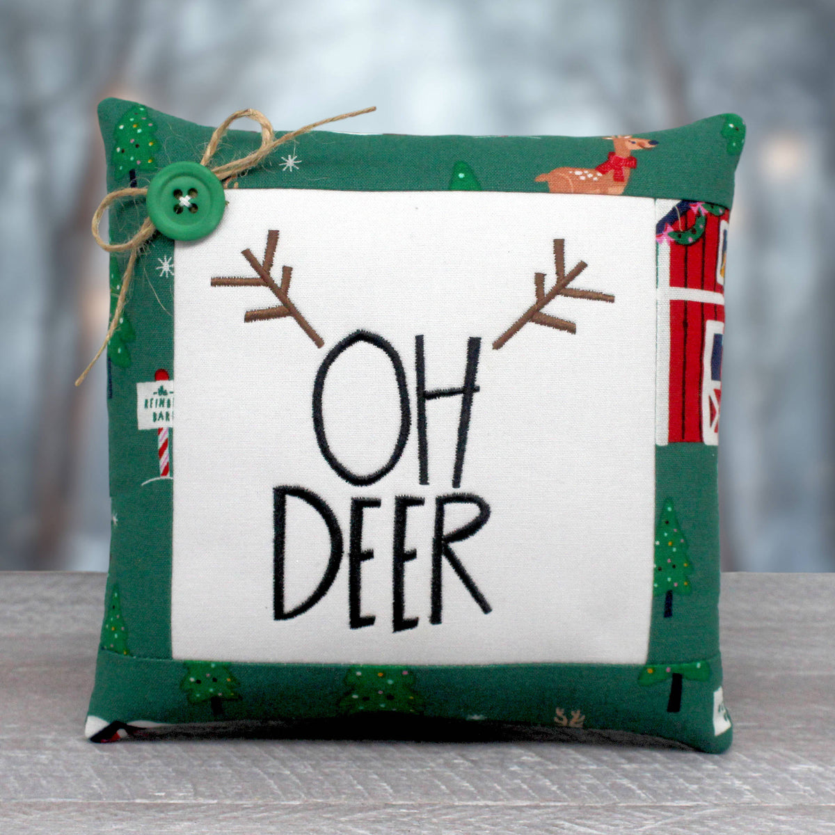 A square decorative throw pillow with a green and white holiday-themed design. The center features a white square panel with the words 'OH DEER' in black embroidery, flanked by brown antler motifs.