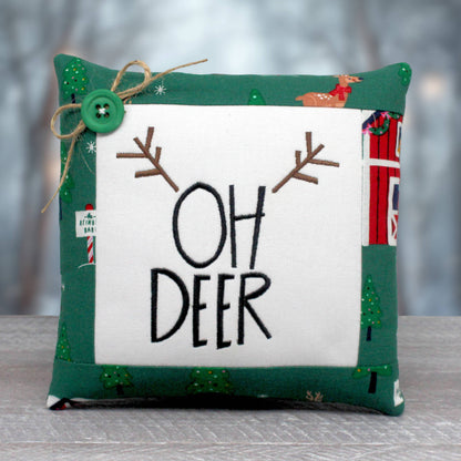 A square decorative throw pillow with a green and white holiday-themed design. The center features a white square panel with the words 'OH DEER' in black embroidery, flanked by brown antler motifs.