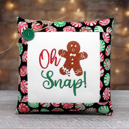 A square decorative throw pillow with a festive holiday design. The center features a white fabric panel with embroidered text reading 'Oh Snap!' in red and green, alongside a brown gingerbread man.