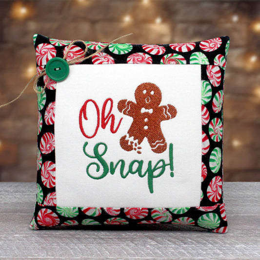A square decorative throw pillow with a festive holiday design. The center features a white fabric panel with embroidered text reading 'Oh Snap!' in red and green, alongside a brown gingerbread man.