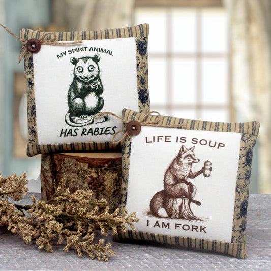 Two decorative fabric mini throw pillows with rustic wood-grain and floral borders, styled on a table with dried flowers and a log. One pillow features an opossum illustration with the text 'MY SPIRIT ANIMAL HAS RABIES', and the other features a fox illustration with the text 'LIFE IS SOUP I AM FORK'.