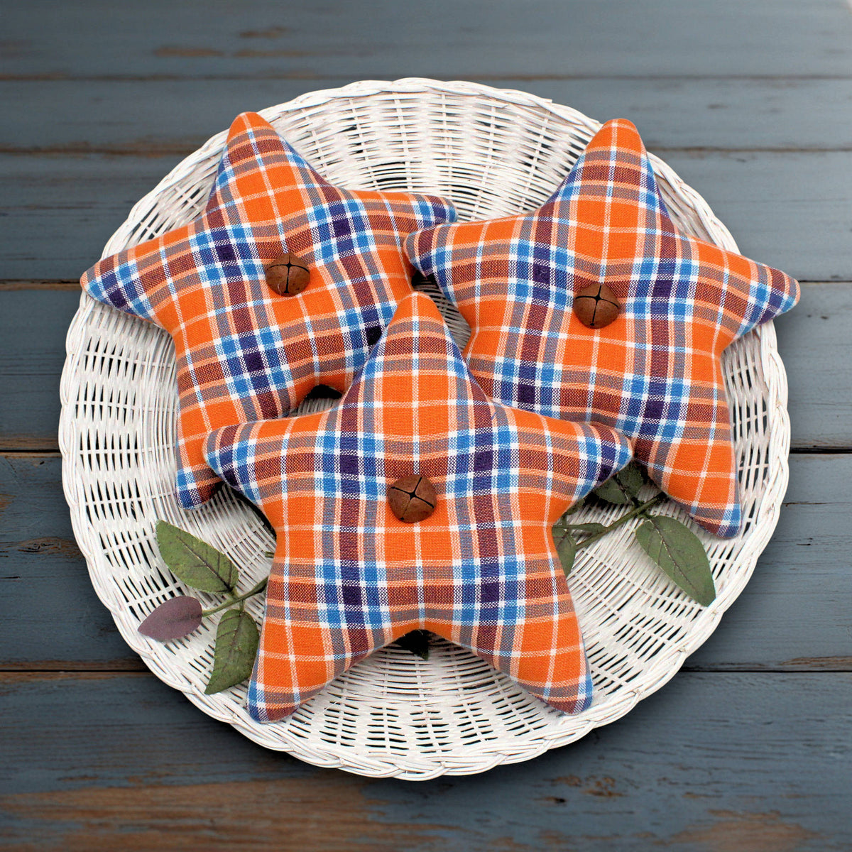 Three star homespun fabric bowl fillers in orange, blue, and white plaid fabric, arranged on a white wicker plate over a dark blue wooden surface.