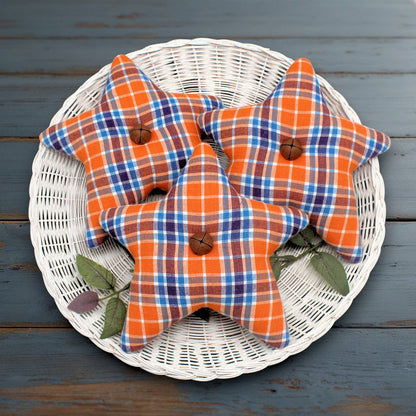 Three star homespun fabric bowl fillers in orange, blue, and white plaid fabric, arranged on a white wicker plate over a dark blue wooden surface.