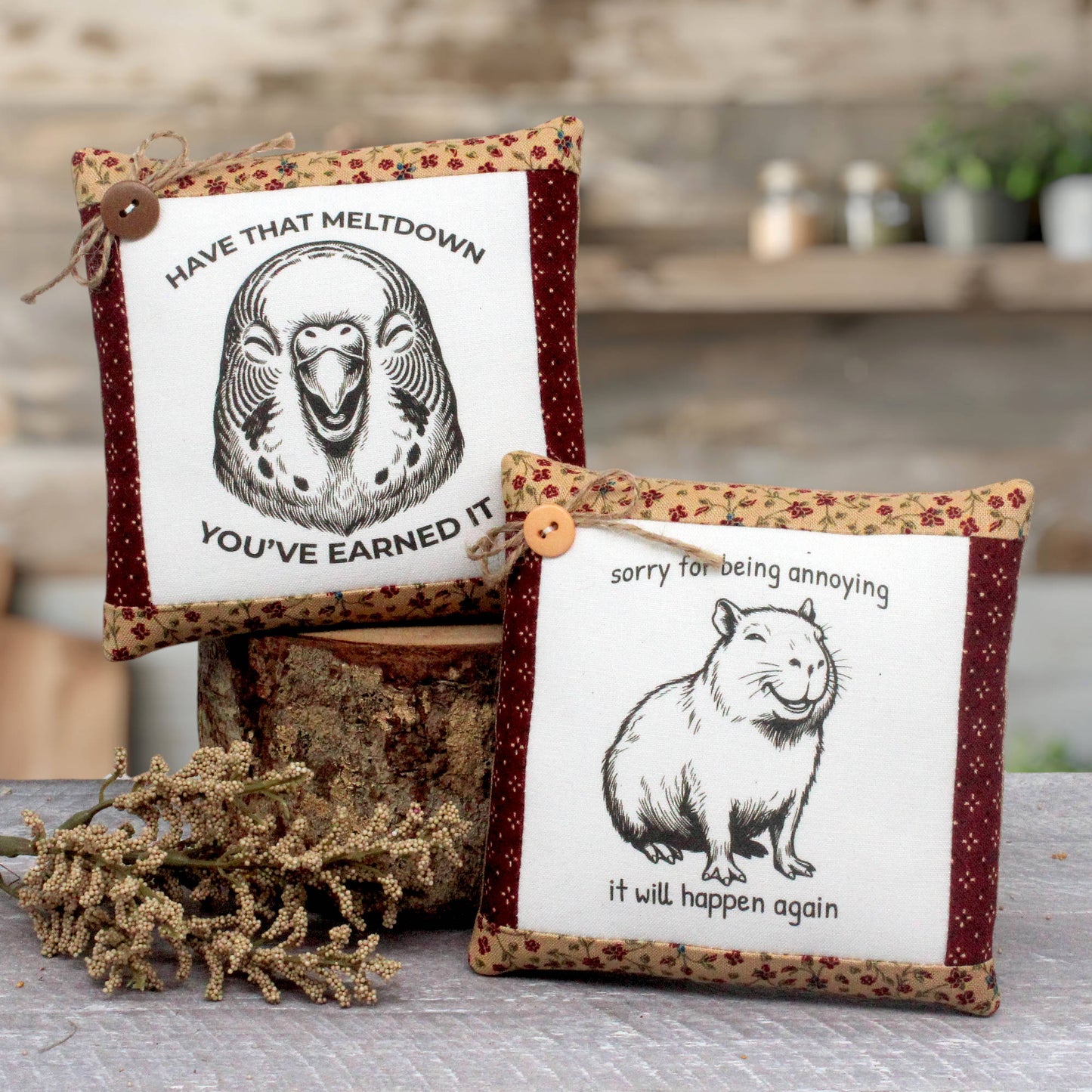 Two decorative square mini pillows with printed animal illustrations and humorous text, styled on a rustic wooden surface with dried flowers and a log in the background.