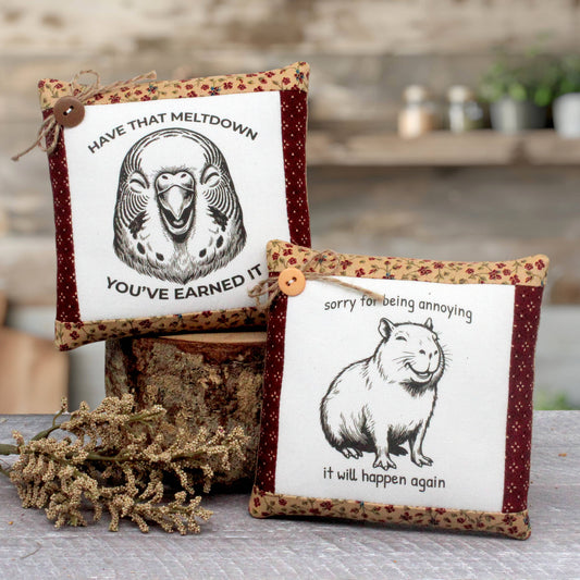 Two decorative square mini pillows with printed animal illustrations and humorous text, styled on a rustic wooden surface with dried flowers and a log in the background.