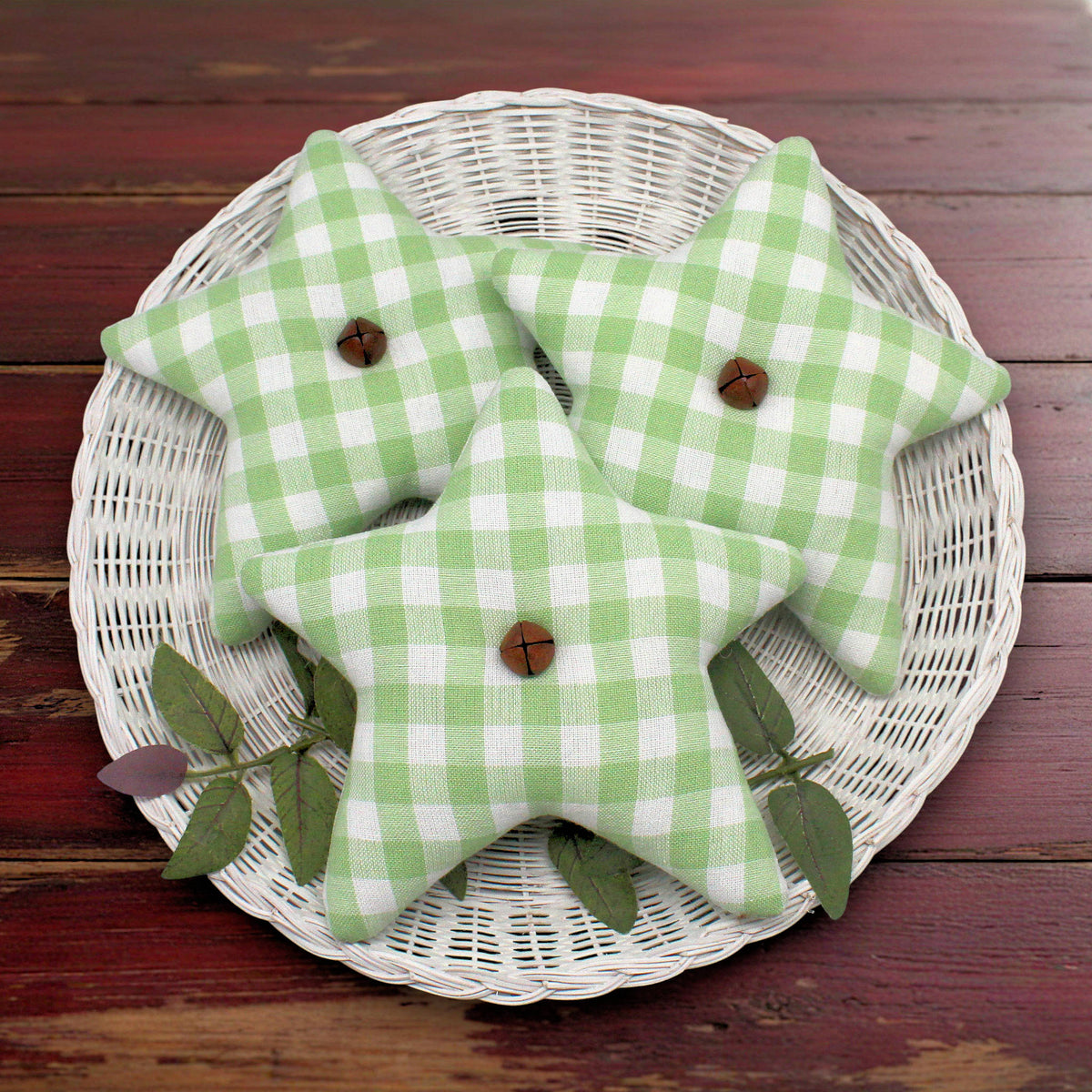 Three homespun fabric stars bowl fillers in a light green and white gingham pattern, arranged on a white wicker tray with green leaves on a dark wooden table.
