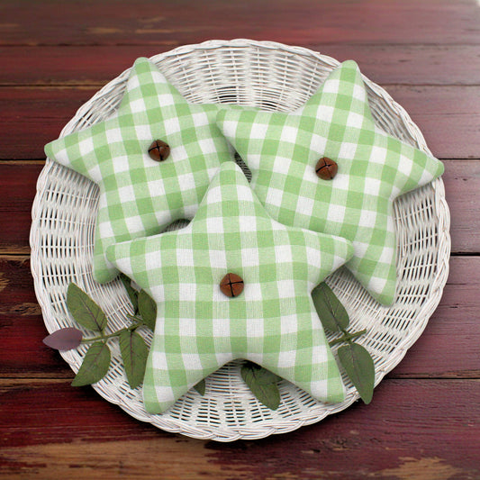 Three homespun fabric stars bowl fillers in a light green and white gingham pattern, arranged on a white wicker tray with green leaves on a dark wooden table.