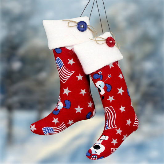 Two red Christmas stockings with white fleece cuffs, decorated with white stars, blue hearts, American flag motifs, and cartoon dog faces, hanging against a blurred snowy outdoor background.