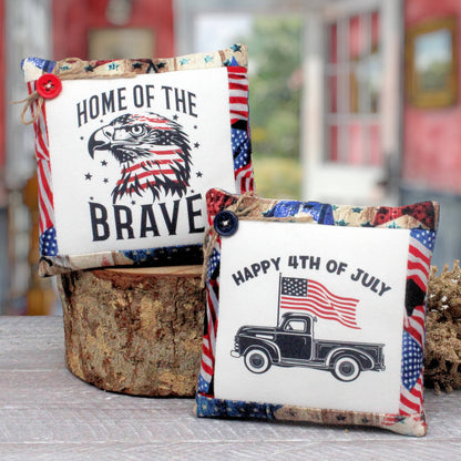 Two patriotic-themed decorative mini pillows displayed on a rustic wood stump. The pillow on the left reads 'HOME OF THE BRAVE' with an eagle graphic in an American flag pattern.