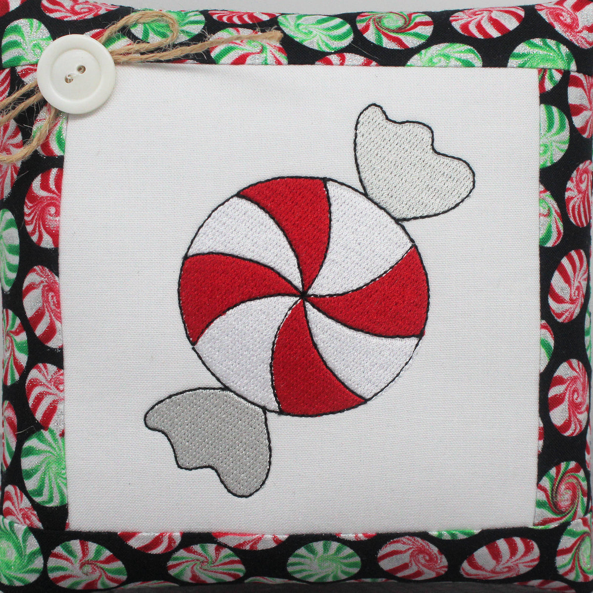 A square mini pillow with an embroidered peppermint candy design with red and white swirls and gray candy tails, bordered by a black fabric with a red, green, and white peppermint pattern. A white button and twine are attached to the top left corner.