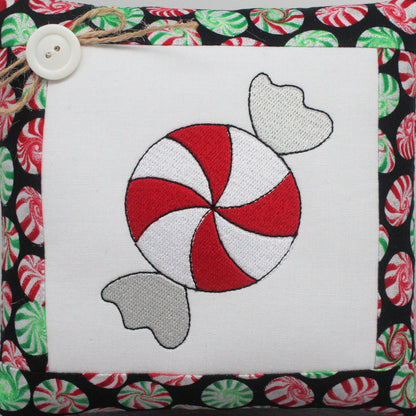 A square mini pillow with an embroidered peppermint candy design with red and white swirls and gray candy tails, bordered by a black fabric with a red, green, and white peppermint pattern. A white button and twine are attached to the top left corner.