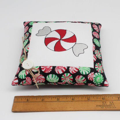 A small square fabric pillow with a Christmas candy design, shown on a white background with a wooden ruler at the bottom for scale. The pillow features a black border with red, green, and white peppermint candy patterns, a white center panel with a red and white swirl candy motif, and a small white button with twine on the side.