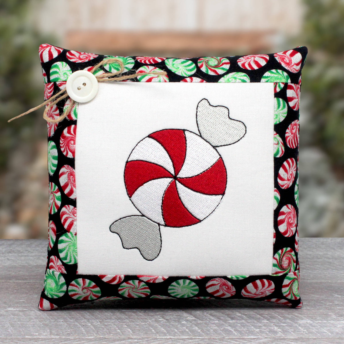 A square decorative throw pillow with a white center panel featuring an embroidered red-and-white peppermint candy motif, surrounded by a black border printed with red, green, and white candy designs. A small white button and twine tie are attached near the top left.