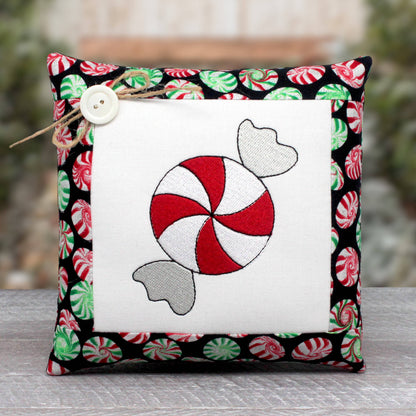A square decorative throw pillow with a white center panel featuring an embroidered red-and-white peppermint candy motif, surrounded by a black border printed with red, green, and white candy designs. A small white button and twine tie are attached near the top left.